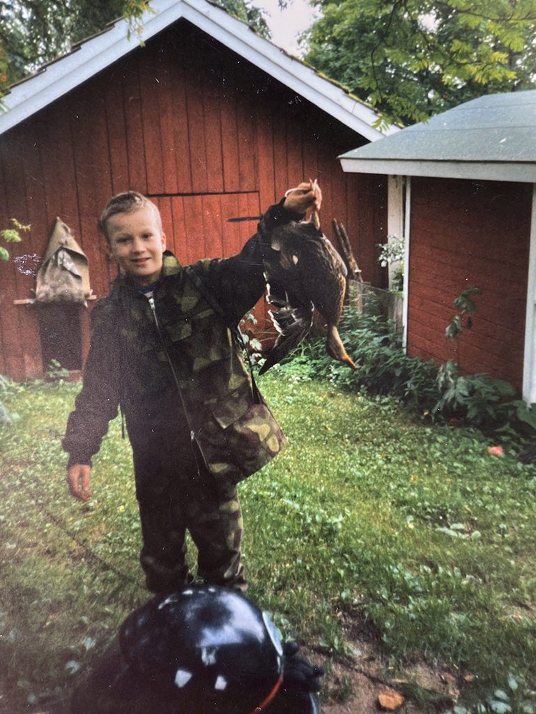 Antti Ruskeala nuorena maastopuvussa punaisen aitan edessä itse ampumansa ensimmäisen heinäsorsan kanssa.